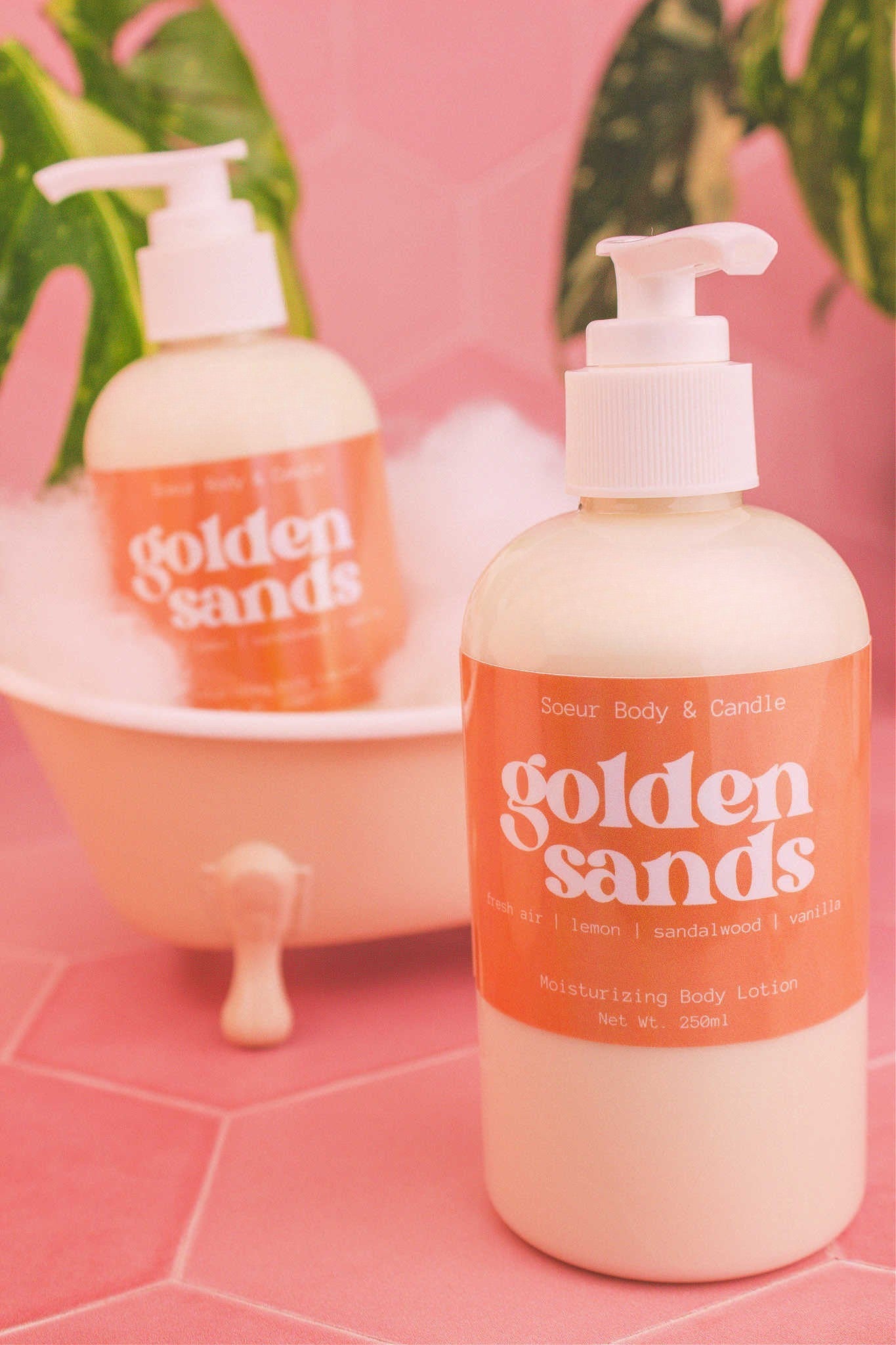 Display of two Golden Sands body lotions in a small tub with bubbles and plants in the background, evoking a relaxing vibe with fresh air, lemon, sandalwood, and vanilla notes.