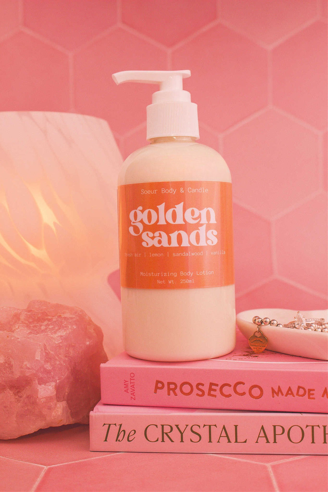 Close-up of the Golden Sands body lotion on a stack of books with a jewelry dish, lamp, and crystal in the background; fragrance notes: fresh air, lemon, sandalwood, and vanilla.