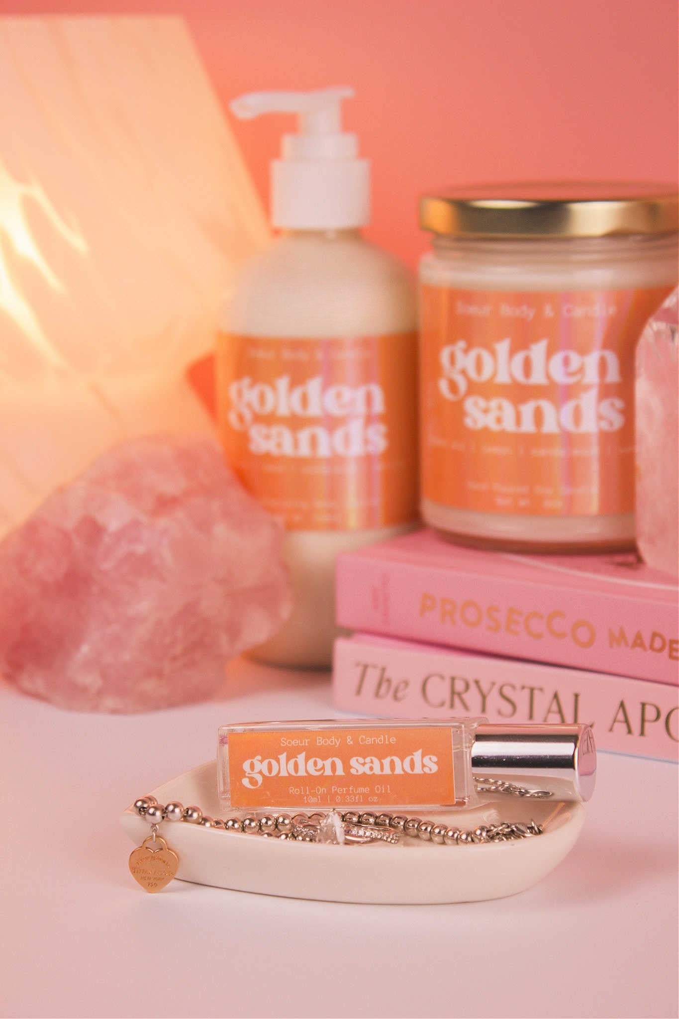 Golden Sands roll-on perfume sitting elegantly in a jewelry dish with jewelry, and coordinating lotion and candle, along with books, a lamp, and crystals are softly blurred in the background. Featuring notes of fresh air, lemon, sandalwood, and vanilla.