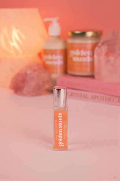 Focused shot of Golden Sands roll-on perfume in the foreground with its matching lotion and candle, along with books, a lamp, and crystals softly blurred in the background. Featuring notes of fresh air, lemon, sandalwood, and vanilla.