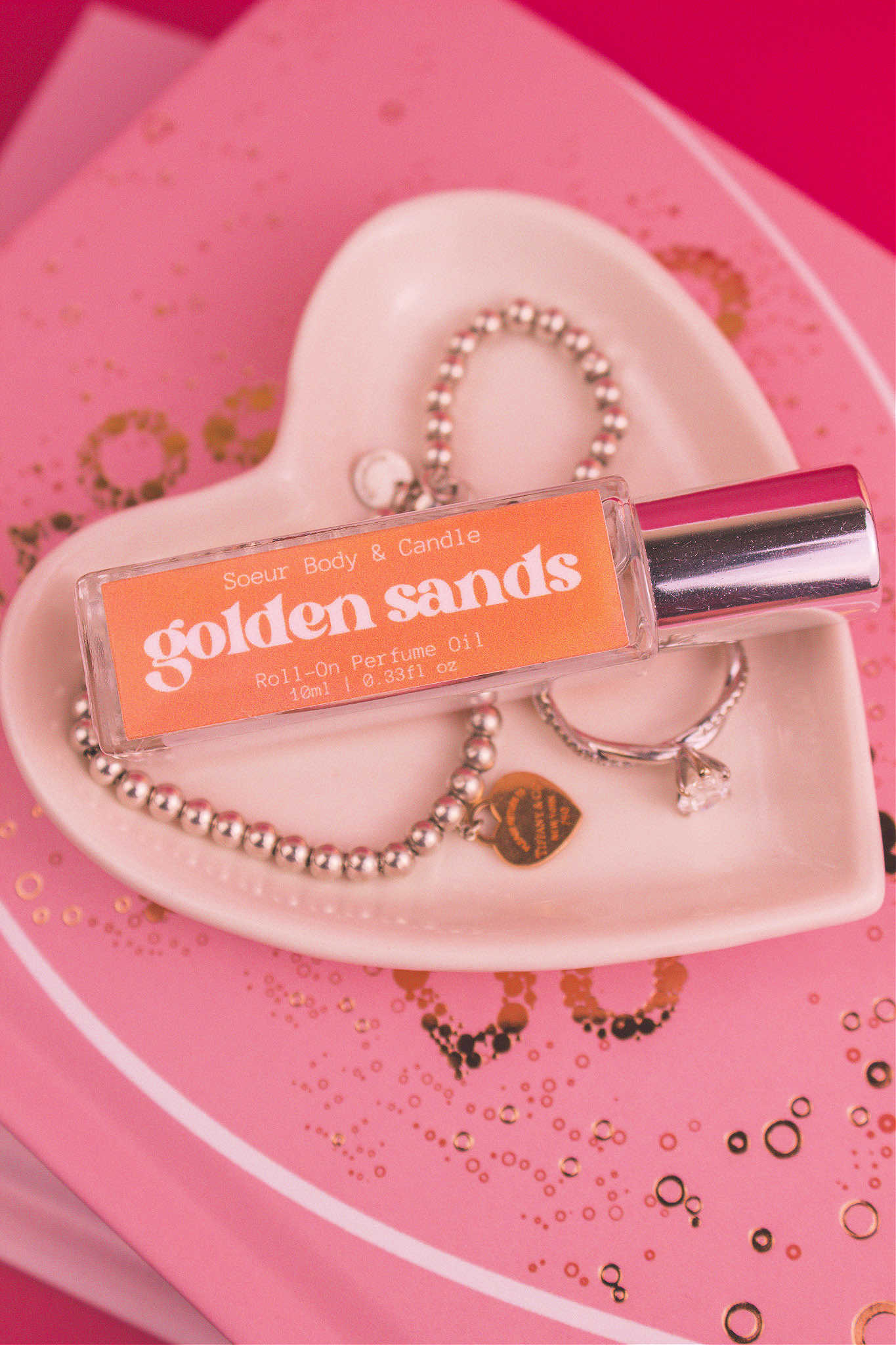 Golden Sands roll-on perfume bottle displayed in a jewelry dish beside a ring and bracelet, sitting on top of books, highlighting its refreshing notes of fresh air, lemon, sandalwood, and vanilla.