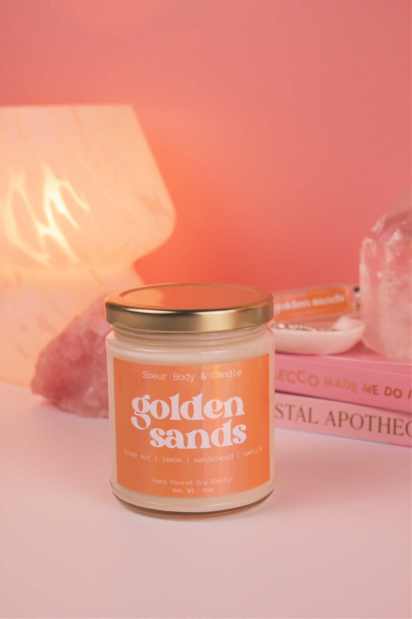 Golden Sands candle with the lid on, with a stack of books and a jewelry dish in the background, set against a lamp and crystals; fragrance notes: fresh air, lemon, sandalwood, and vanilla.