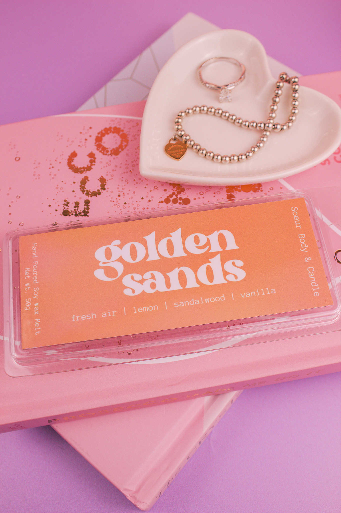 Golden Sands wax melt on a stack of books with a jewelry dish in the background holding jewelry; fragrance notes: fresh air, lemon, sandalwood, and vanilla.