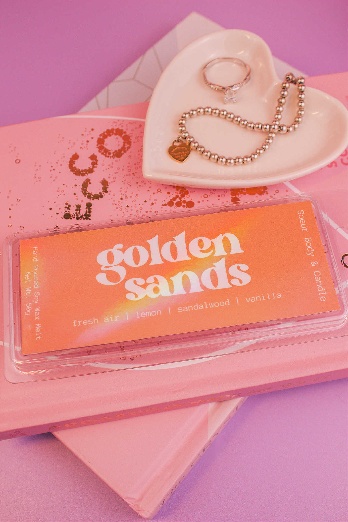 Close-up of the Golden Sands wax melt on a stack of books with a jewelry dish in the background holding jewelry; fragrance notes: fresh air, lemon, sandalwood, and vanilla.