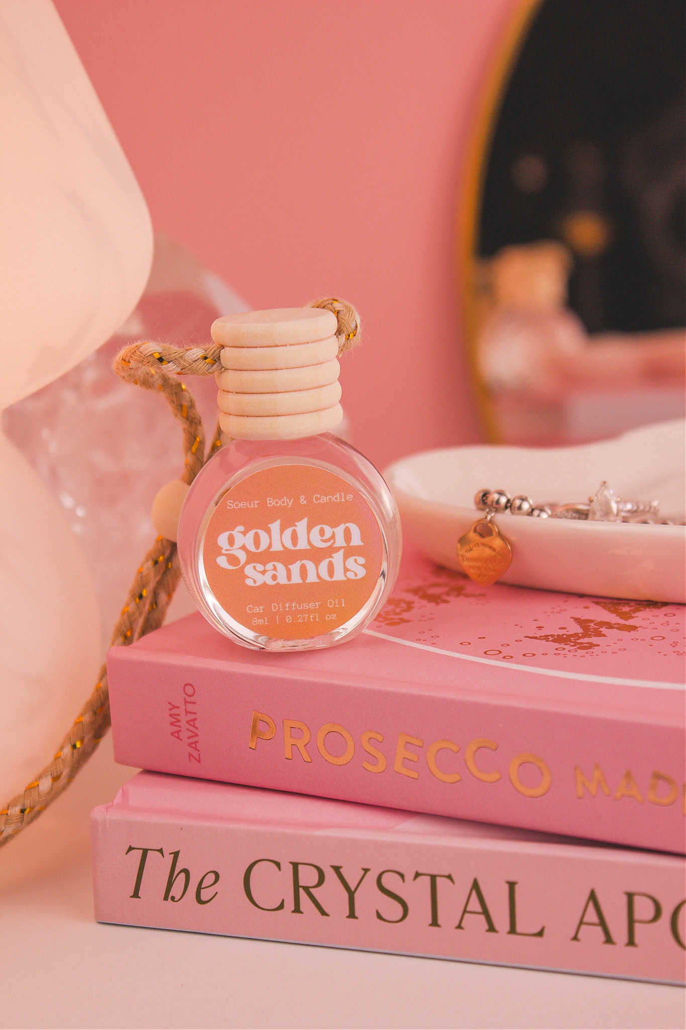 Golden Sands diffuser sitting on top of a stack of books with a jewelry dish in the background, set against a lamp, mirror, and crystals; fragrance notes: fresh air, lemon, sandalwood, and vanilla notes.