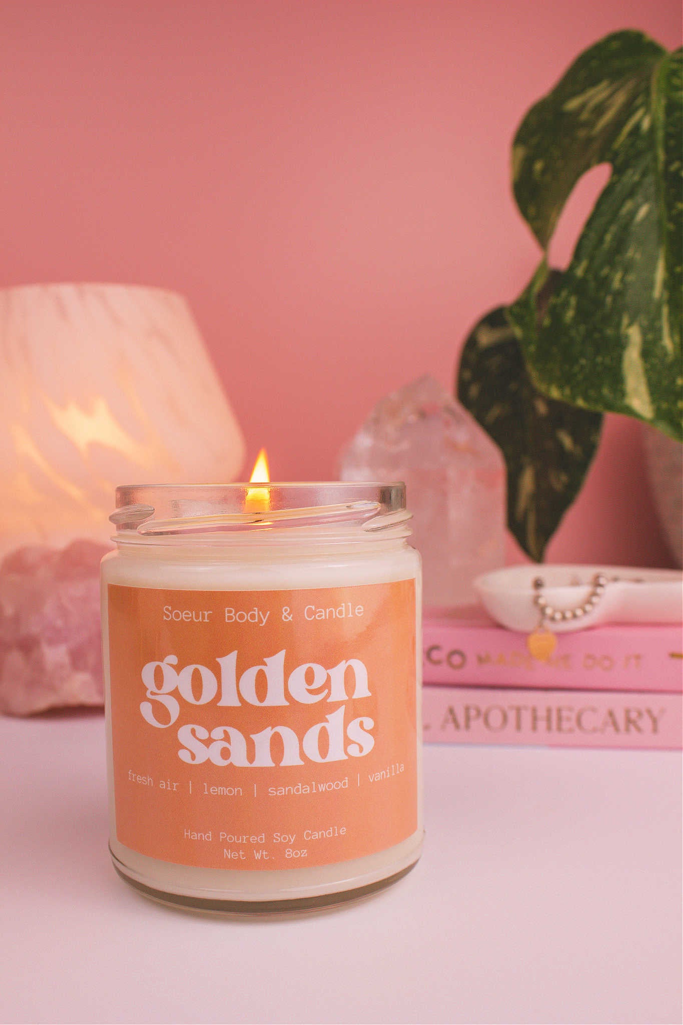 Golden Sands candle lit, with a stack of books and a jewelry dish in the background, set against a lamp, plant, and crystals; fragrance notes: fresh air, lemon, sandalwood, and vanilla.