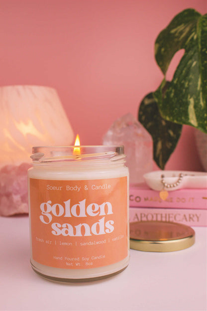 Close-up of Golden Sands candle lit, with a stack of books and a jewelry dish in the background, set against a lamp, plant, and crystals; fragrance notes: fresh air, lemon, sandalwood, and vanilla.