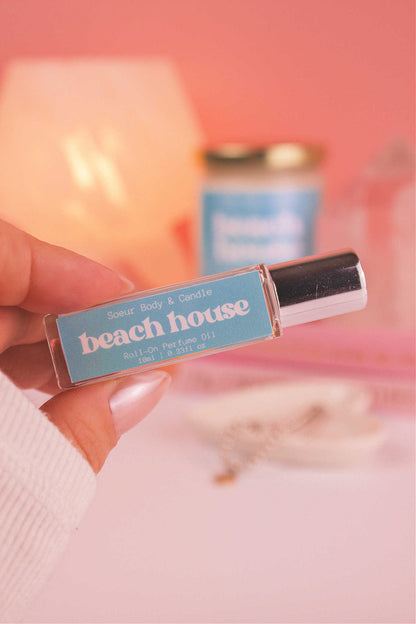 Close-up of Beach House roll-on perfume held in hand with matching candle blurred in the background, showcasing its refreshing blend of citrus fruits, coconut, vanilla, and warm musk.