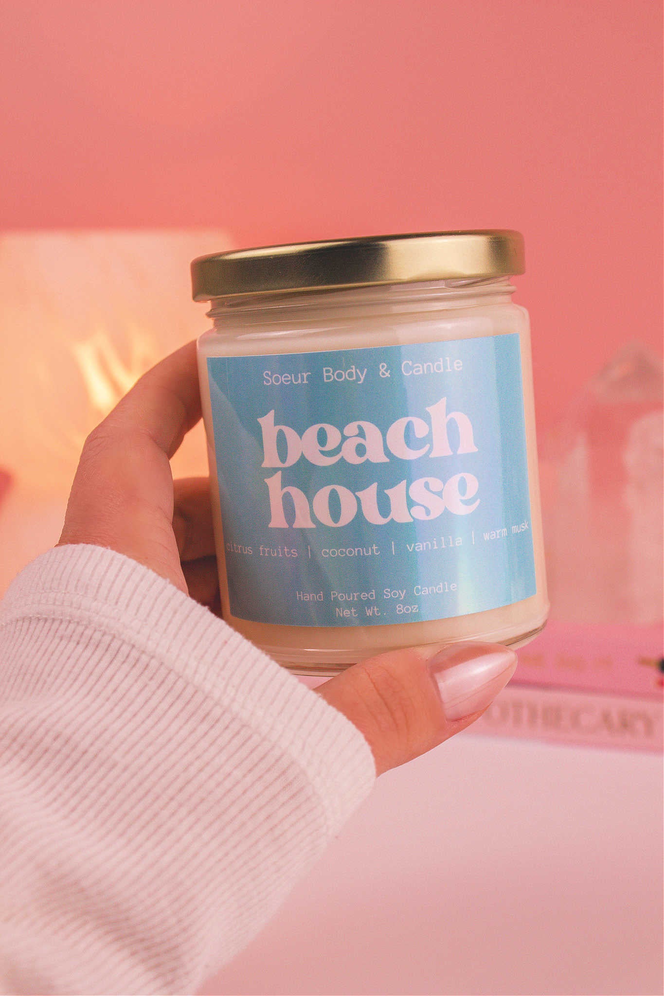 Close-up of a hand holding the Beach House candle, showcasing its design and fragrance notes of citrus fruits, coconut, vanilla, and warm musk.