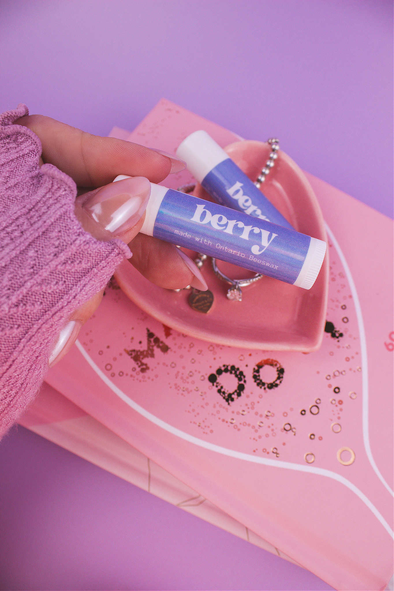 Berry lip balm held in hand with matching lip balm, ring, and bracelet displayed in a jewelry dish on top of books in the background.