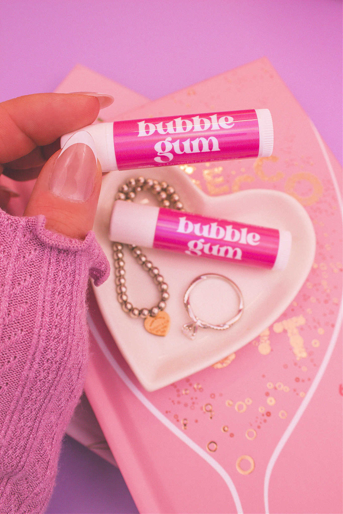 Bubblegum lip balm held in hand with matching lip balm, ring, and bracelet displayed in a jewelry dish on top of books in the background.