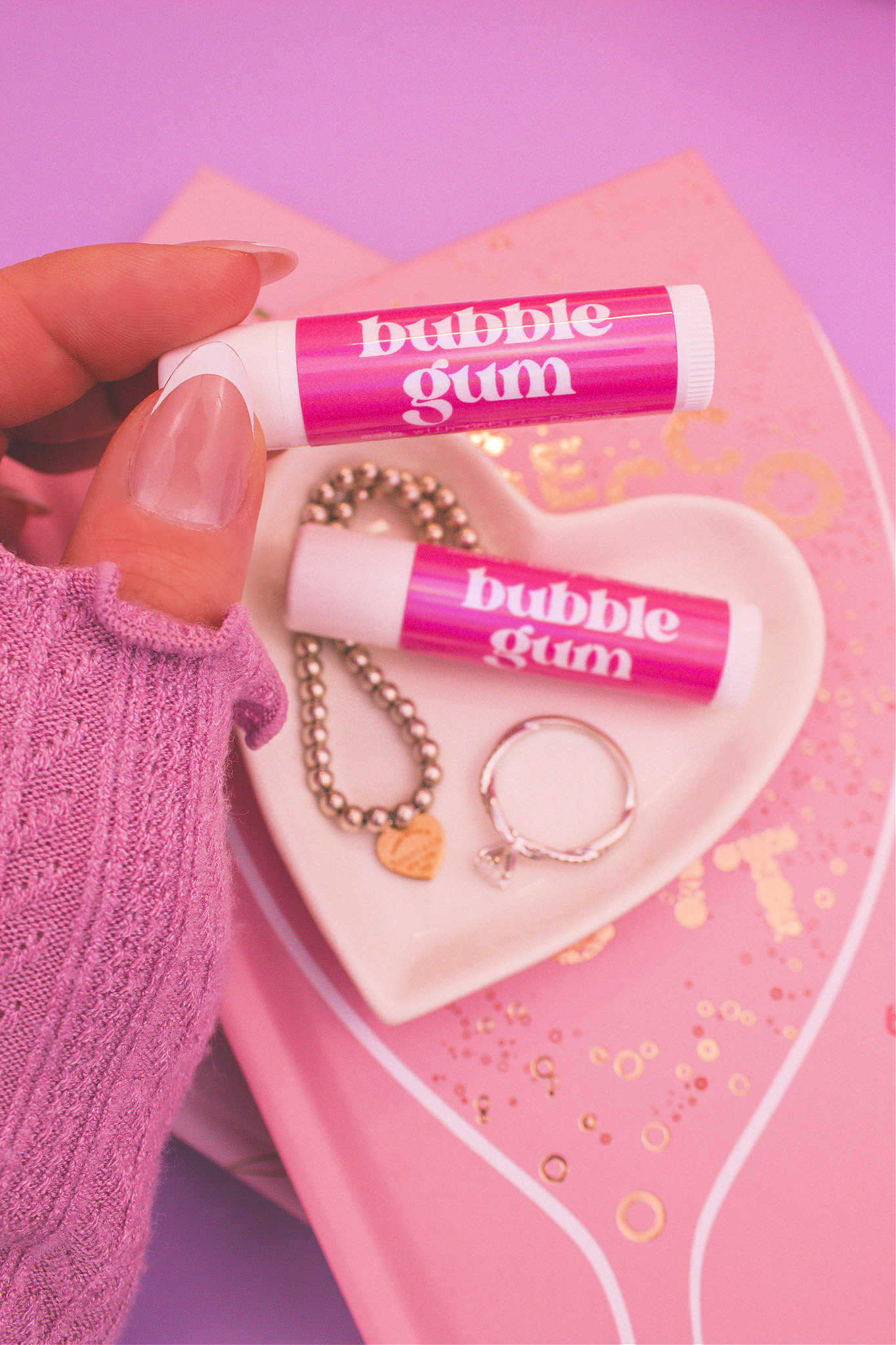 Bubblegum lip balm held in hand with matching lip balm, ring, and bracelet displayed in a jewelry dish on top of books in the background.