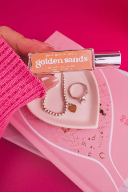 Close-up shot of Golden Sands roll-on perfume held in hand with a jewelry dish and jewelry in the background, capturing its invigorating mix of fresh air, lemon, sandalwood, and vanilla.