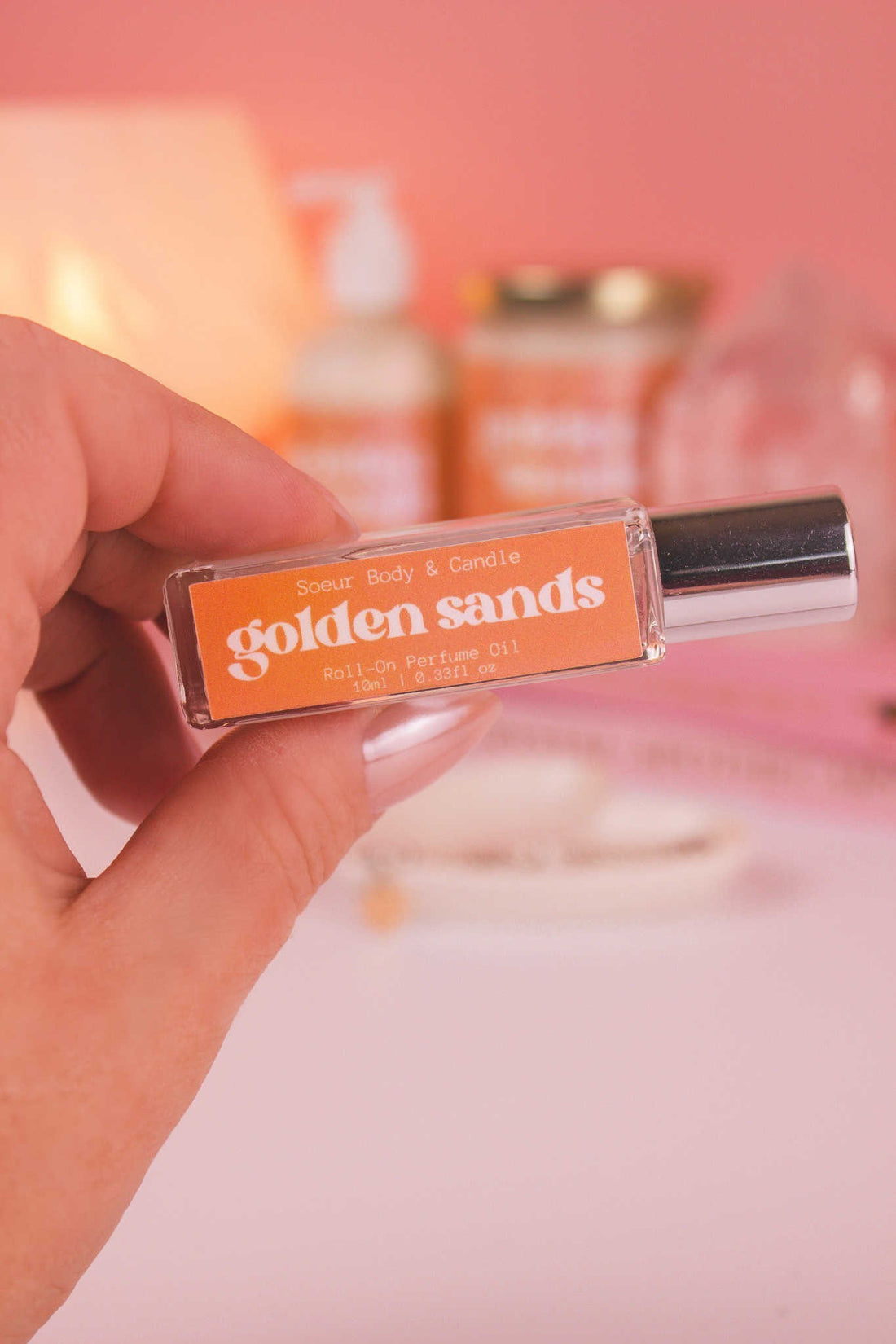 Close-up of Golden Sands roll-on perfume held in hand with matching lotion and candle blurred in the background, showcasing its blend of fresh air, lemon, sandalwood, and vanilla notes.