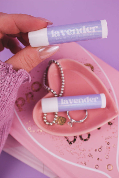 Lavender lip balm held in hand with matching lip balm, ring, and bracelet displayed in a jewelry dish on top of books in the background.