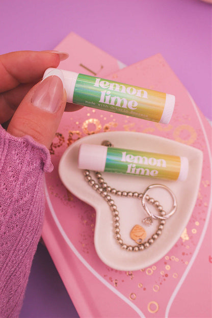 Lemon Lime lip balm held in hand with matching lip balm, ring, and bracelet displayed in a jewelry dish on top of books in the background.