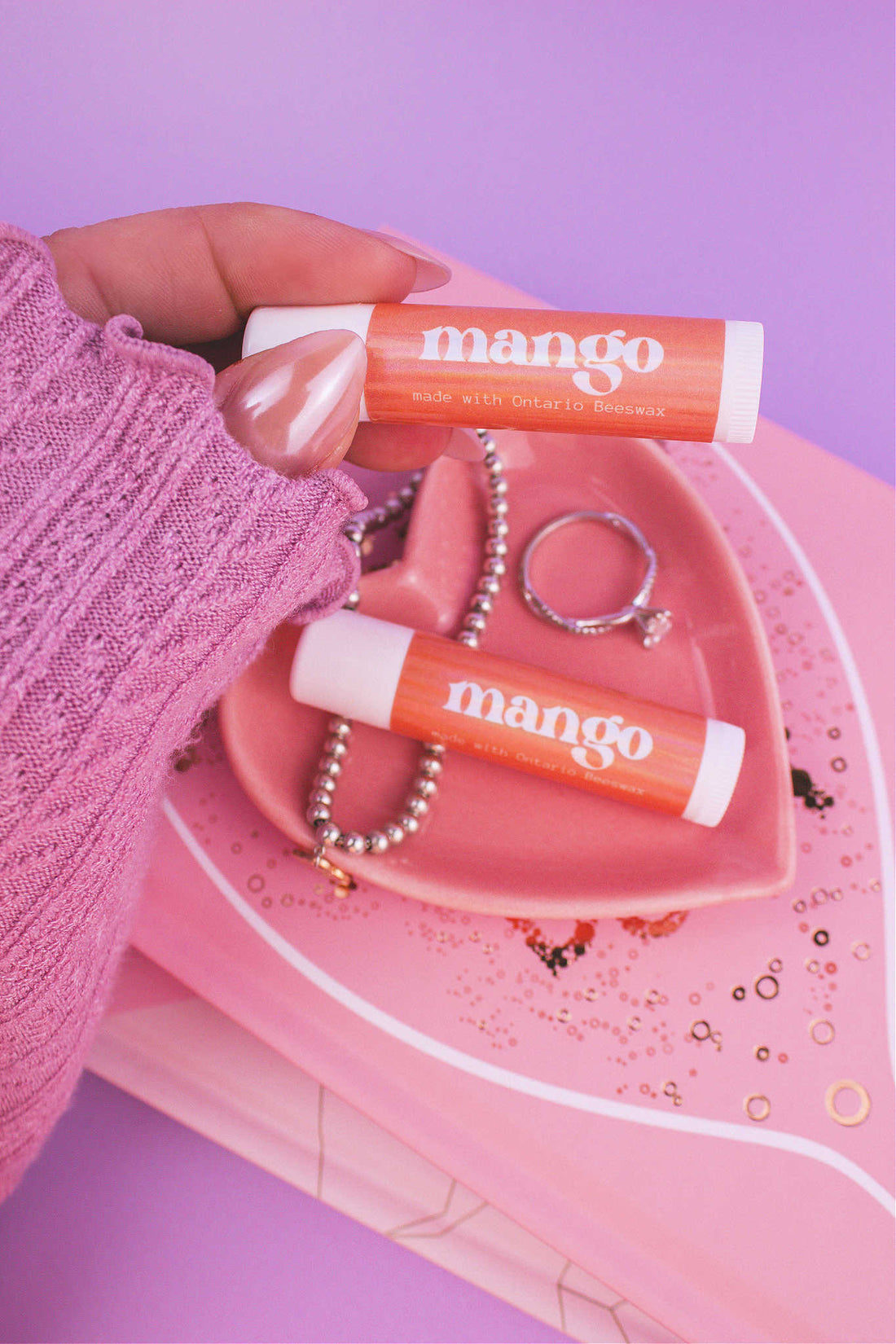 Mango lip balm held in hand with matching lip balm, ring, and bracelet displayed in a jewelry dish on top of books in the background.