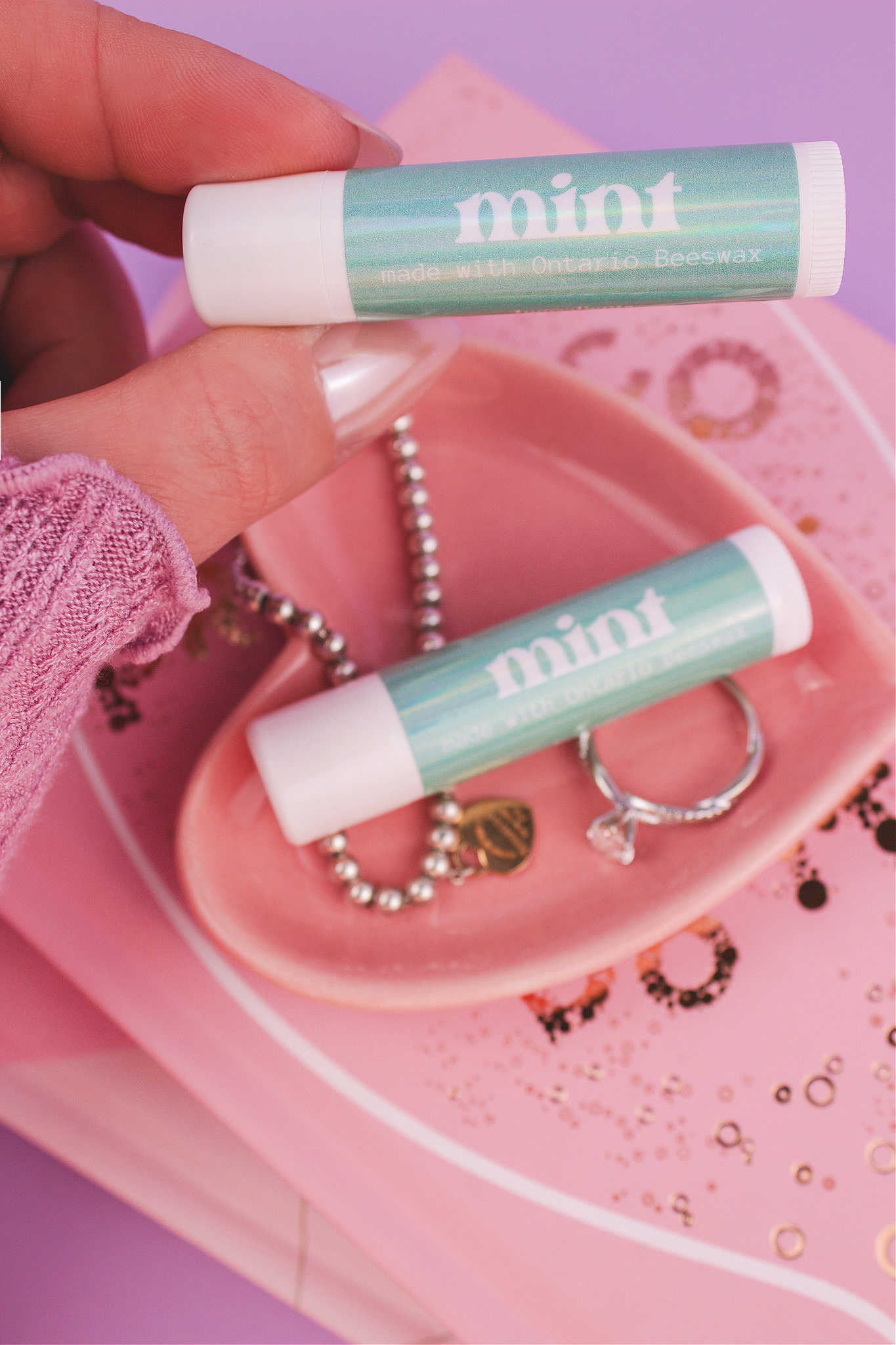 Mint lip balm held in hand with matching lip balm, ring, and bracelet displayed in a jewelry dish on top of books in the background.