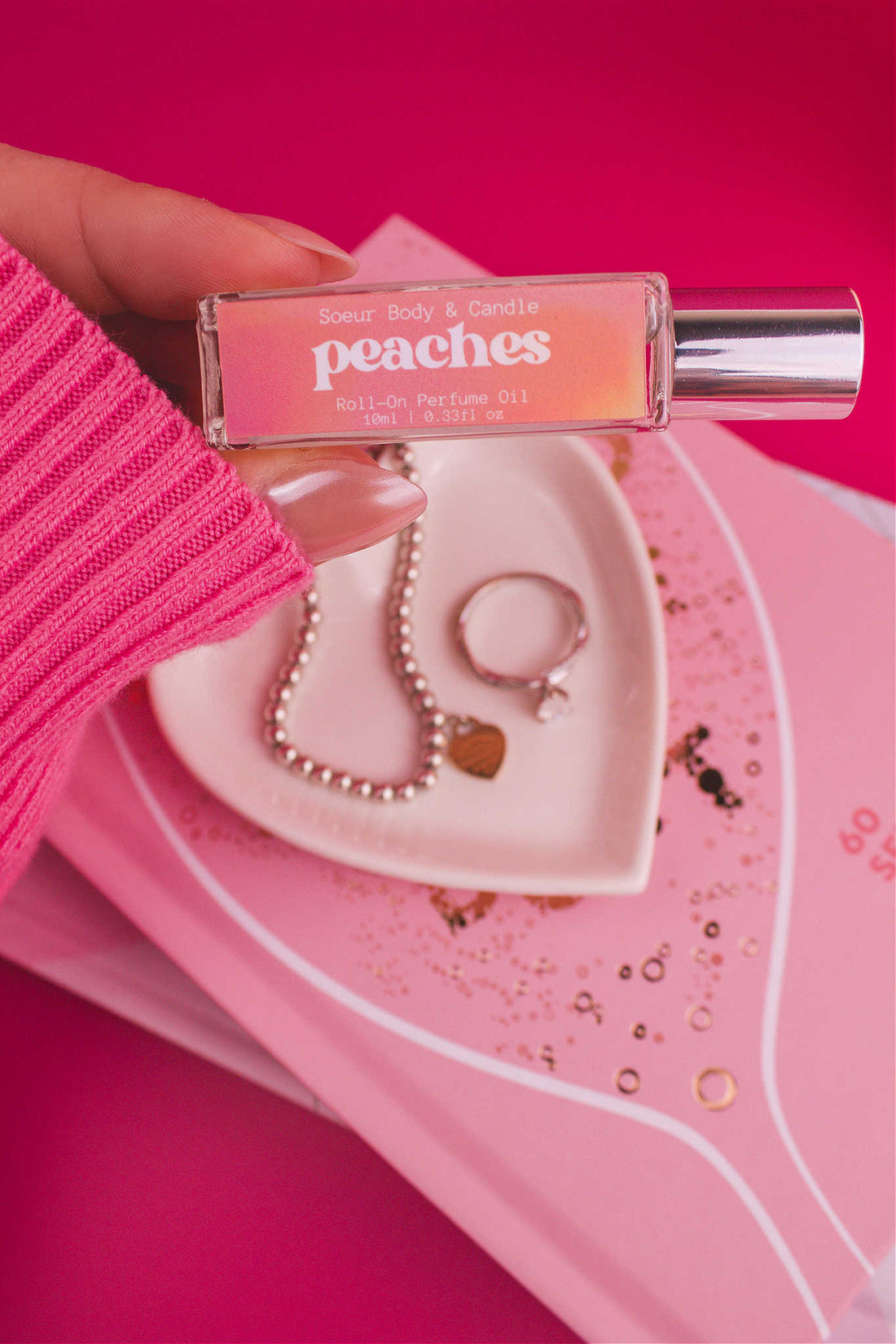 Close-up of Peaches roll-on perfume held in hand with jewelry dish and books in the background, featuring a sweet blend of orange, persimmon, jasmine, peach, and musk.