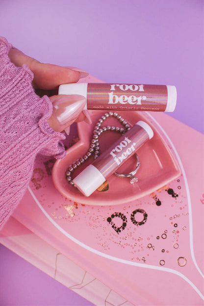 Root Beer lip balm held in hand with matching lip balm, ring, and bracelet displayed in a jewelry dish on top of books in the background.