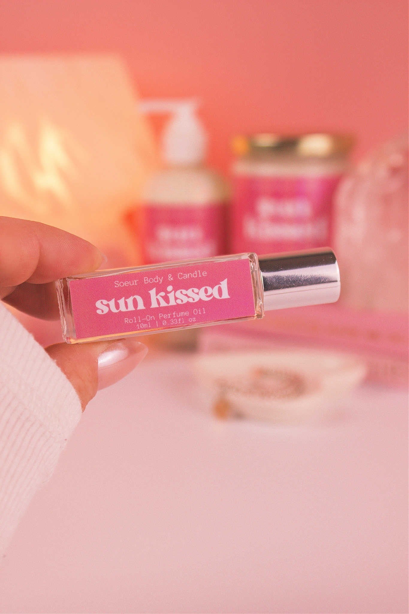 Close-up of Sun Kissed roll-on perfume held in hand with matching lotion and candle in the background, featuring notes of salted caramel, pistachio, almond, and vanilla.