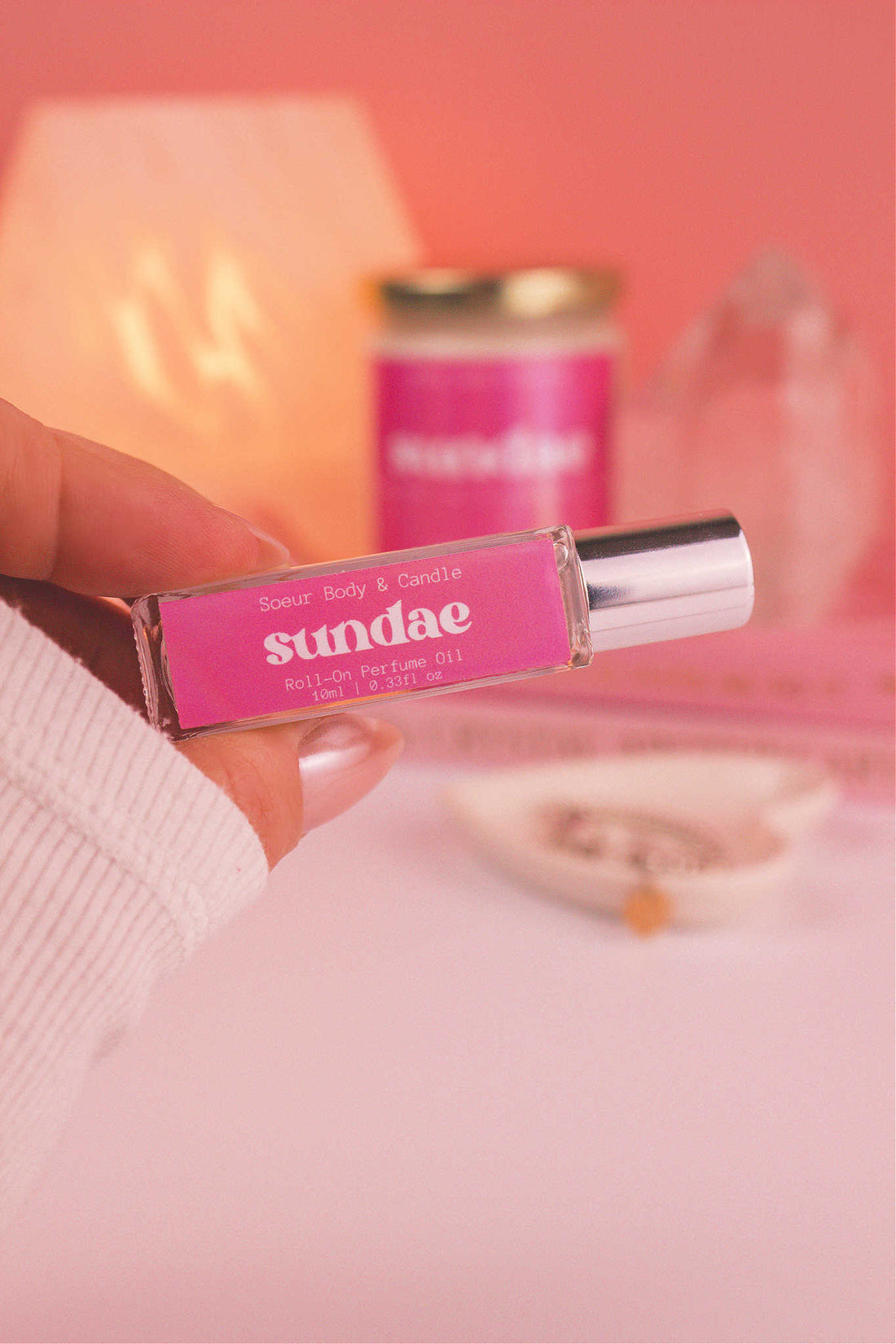 Close-up of Sundae roll-on perfume held in hand with matching candle in the background, showcasing a sweet blend of sweet cream, vanilla, and sugary waffle cone.