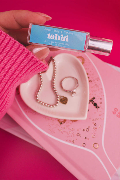 Close-up shot of Tahiti roll-on perfume held in hand with a jewelry dish and jewelry in the background, capturing its vibrant mix of sugar, vanilla, almond, amber, and oak moss notes.