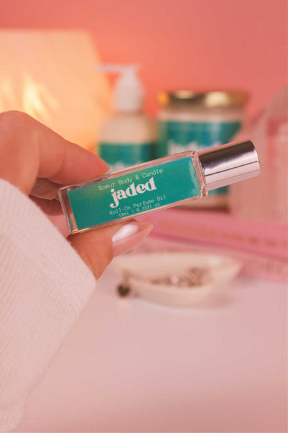 Close-up of Jaded roll-on perfume held in hand with matching lotion and candle in the background, showcasing its complex blend of spruce, pine, amber, vetiver, and musk.