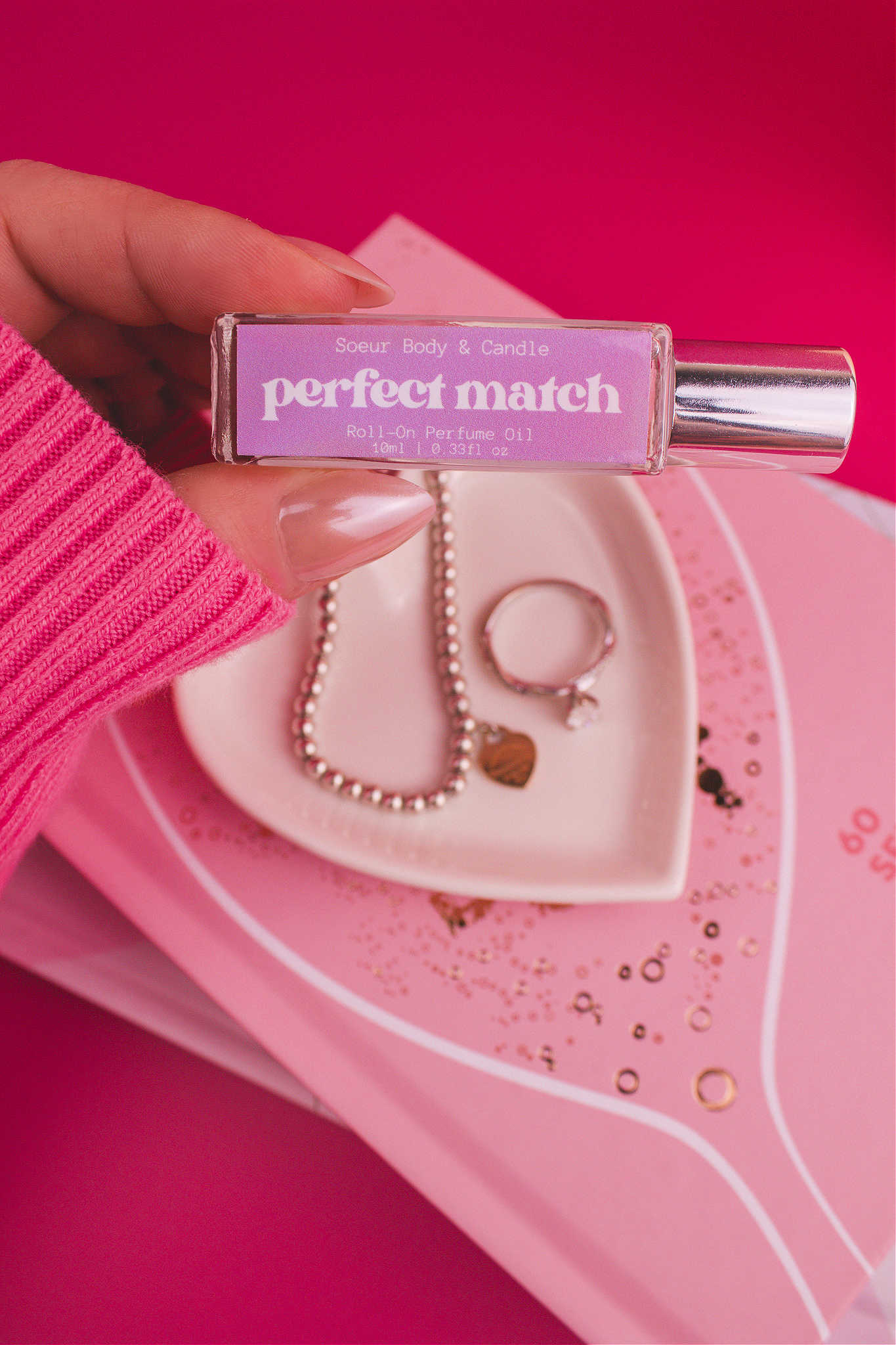 Close-up shot of Perfect Match roll-on perfume held in hand with a jewelry dish and jewelry in the background, capturing its complex blend of cherry, almond, jasmine, rose, and vanilla.