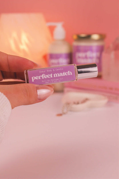 Close-up shot of Perfect Match roll-on perfume held in hand, with a jewelry dish, matching candle and lotion, books, a lamp, and crystals in the background. Featuring notes of cherry, almond, jasmine, rose, and vanilla.