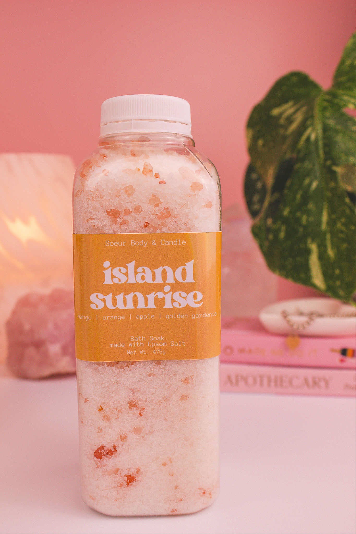 Island Sunrise bath soak with a stack of books, a jewelry dish, lamp, plants, and crystals in the background. Fragrance notes: mango, orange, apple, and golden gardenia.