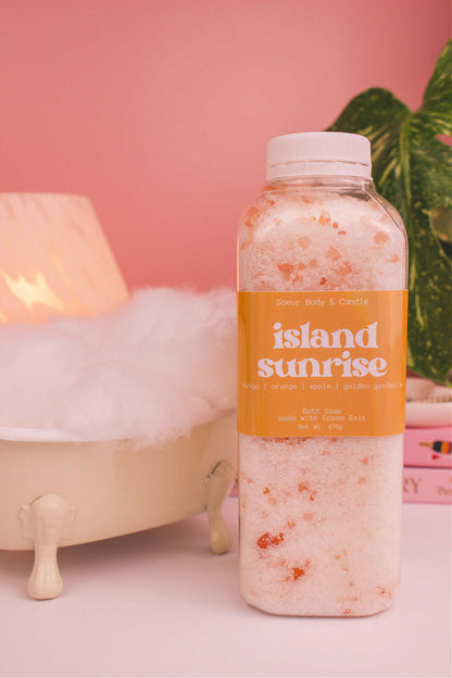 Island Sunrise bath soak beside a small tub with bubbles, a lamp, and plants in the background, creating a relaxing vibe. Fragrance notes: mango, orange, apple, and golden gardenia.