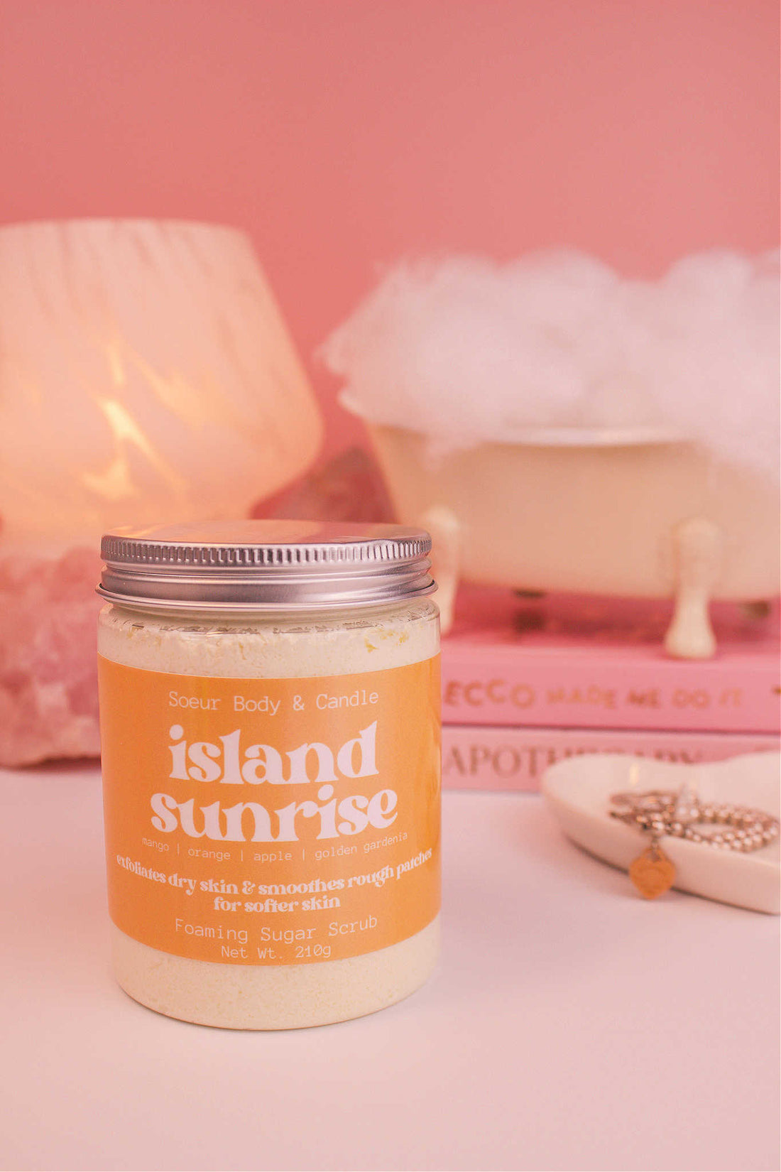 Island Sunrise sugar scrub in the foreground, with a small tub filled with bubbles sitting atop a stack of books alongside a jewelry dish, lamp, and crystals. Fragrance notes: mango, orange, apple, and golden gardenia.
