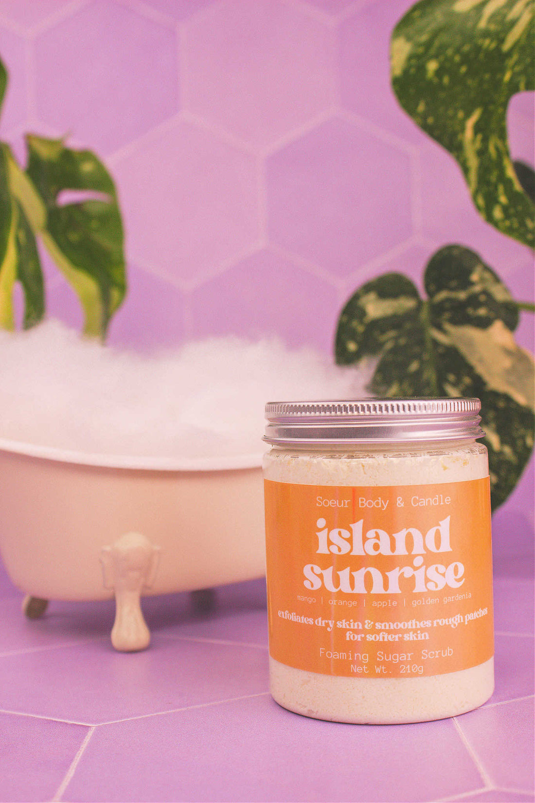 Island Sunrise sugar scrub in front of a small tub with bubbles, and plants in the background, evoking a relaxing vibe with mango, orange, apple, and golden gardenia notes.