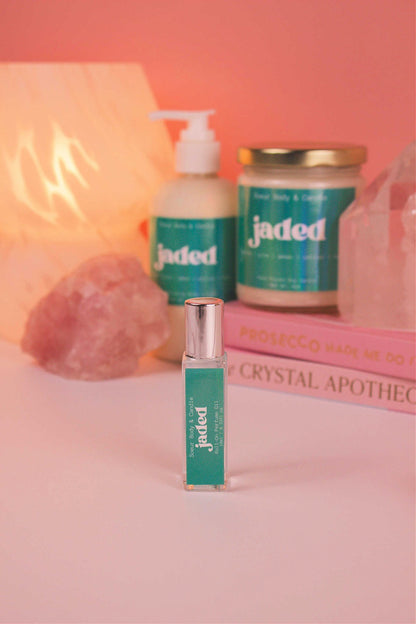 Focused shot of Jaded roll-on perfume in the foreground with its matching lotion and candle, along with books, a lamp, and crystals softly blurred in the background. Featuring notes of spruce, pine, amber, vetiver, and musk.