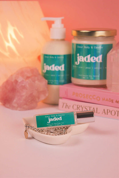 Jaded roll-on perfume elegantly set in a jewelry dish with jewelry, and coordinating lotion and candle, along with books, a lamp, and crystals in the background. Featuring notes of spruce, pine, amber, vetiver, and musk.