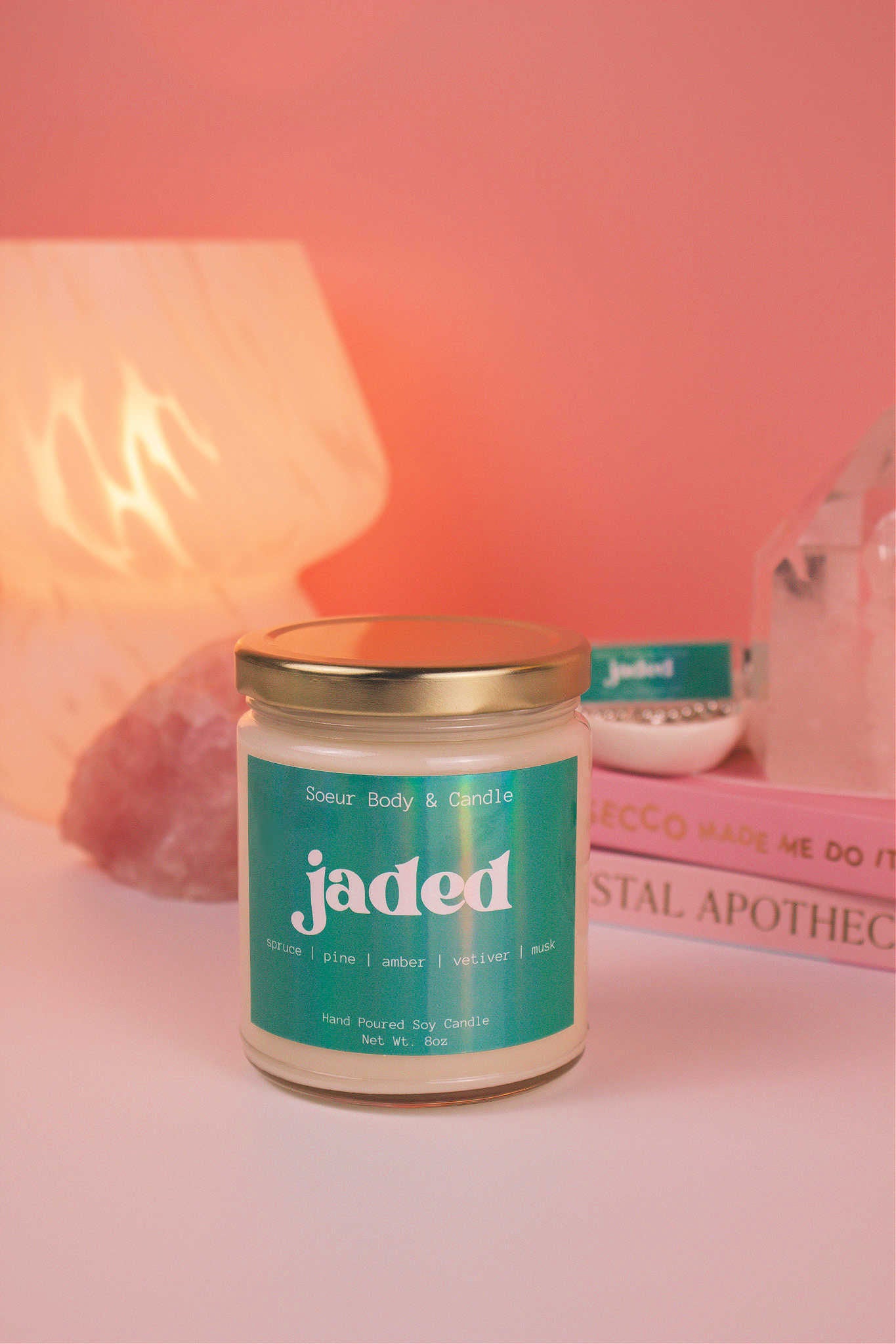 Close-up of Jaded candle with the lid on, with a stack of books and a jewelry dish in the background, set against a lamp and crystals; fragrance notes: spruce, pine, amber, vetiver, and musk.