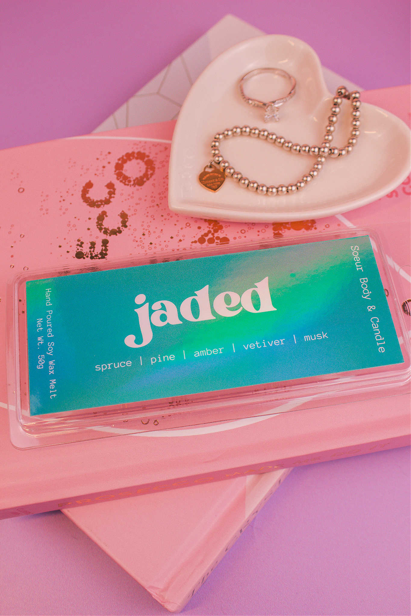 Close-up of the Jaded wax melt on top of books with a jewelry dish in the background displaying jewelry; fragrance notes: spruce, pine, amber, vetiver, and musk.