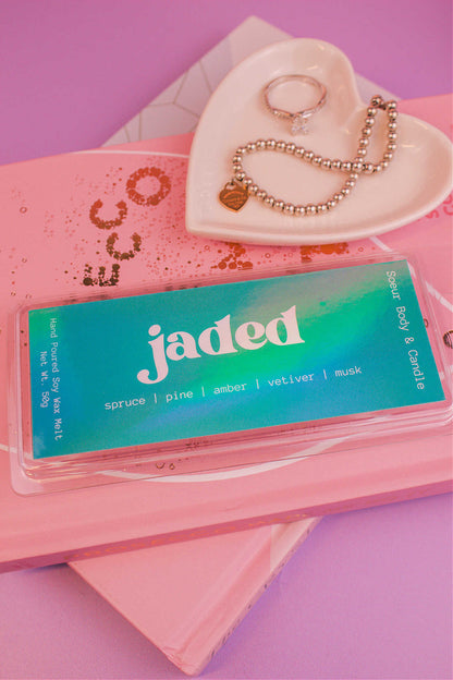 Close-up of the Jaded wax melt on top of books with a jewelry dish in the background displaying jewelry; fragrance notes: spruce, pine, amber, vetiver, and musk.