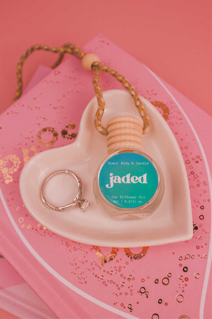 Jaded car diffuser in a jewelry dish with a ring beside it, sitting on top of books, featuring a complex blend of spruce, pine, amber, vetiver, and musk.