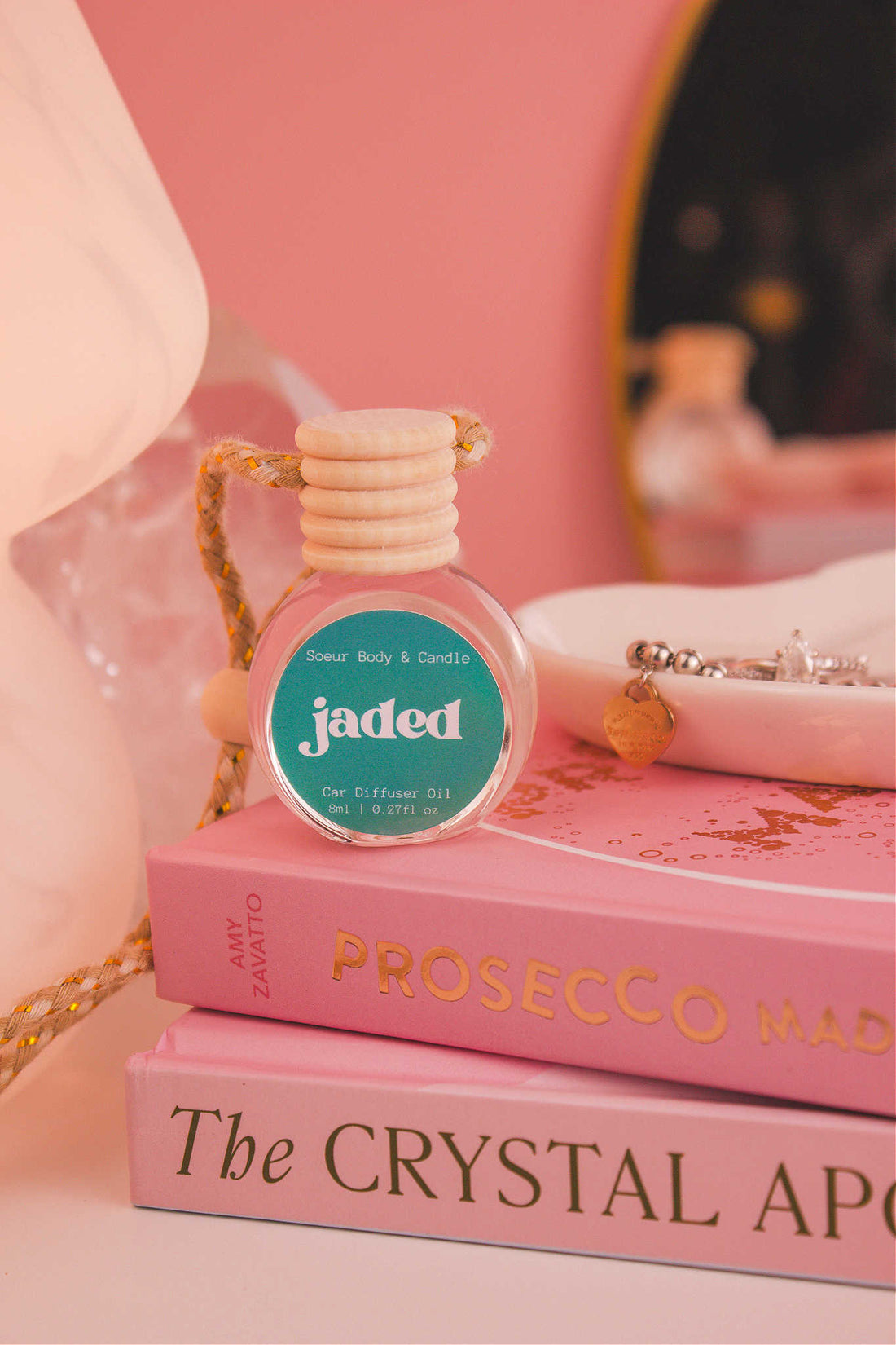 Jaded diffuser sitting on top of a stack of books with a jewelry dish in the background, set against a lamp, mirror, and crystals; fragrance notes: spruce, pine, amber, vetiver, and musk.