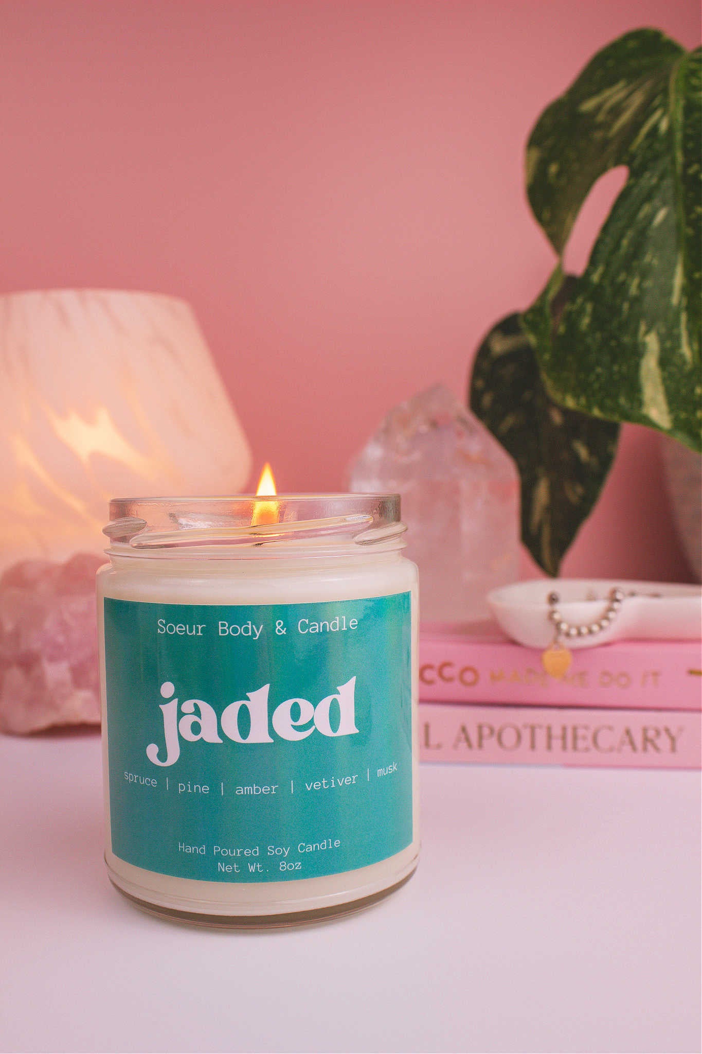 Jaded candle lit, with a stack of books and a jewelry dish in the background, set against a lamp, plant, and crystals; fragrance notes: spruce, pine, amber, vetiver, and musk.