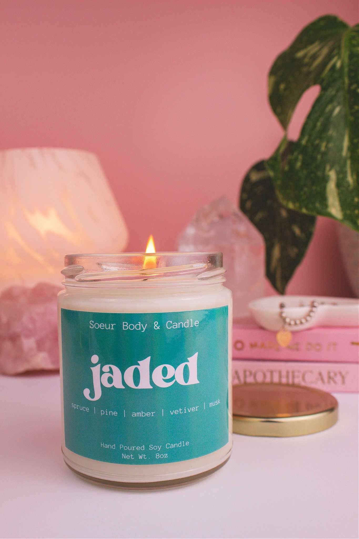 Close-up of Jaded candle lit, with a stack of books and a jewelry dish in the background, set against a lamp, plant, and crystals; fragrance notes: spruce, pine, amber, vetiver, and musk.