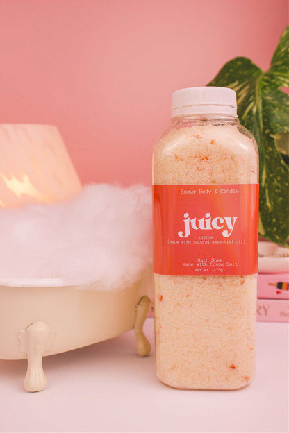 Juicy bath soak beside a small tub with bubbles, a lamp, and plants in the background, creating a relaxing vibe with orange essential oil.