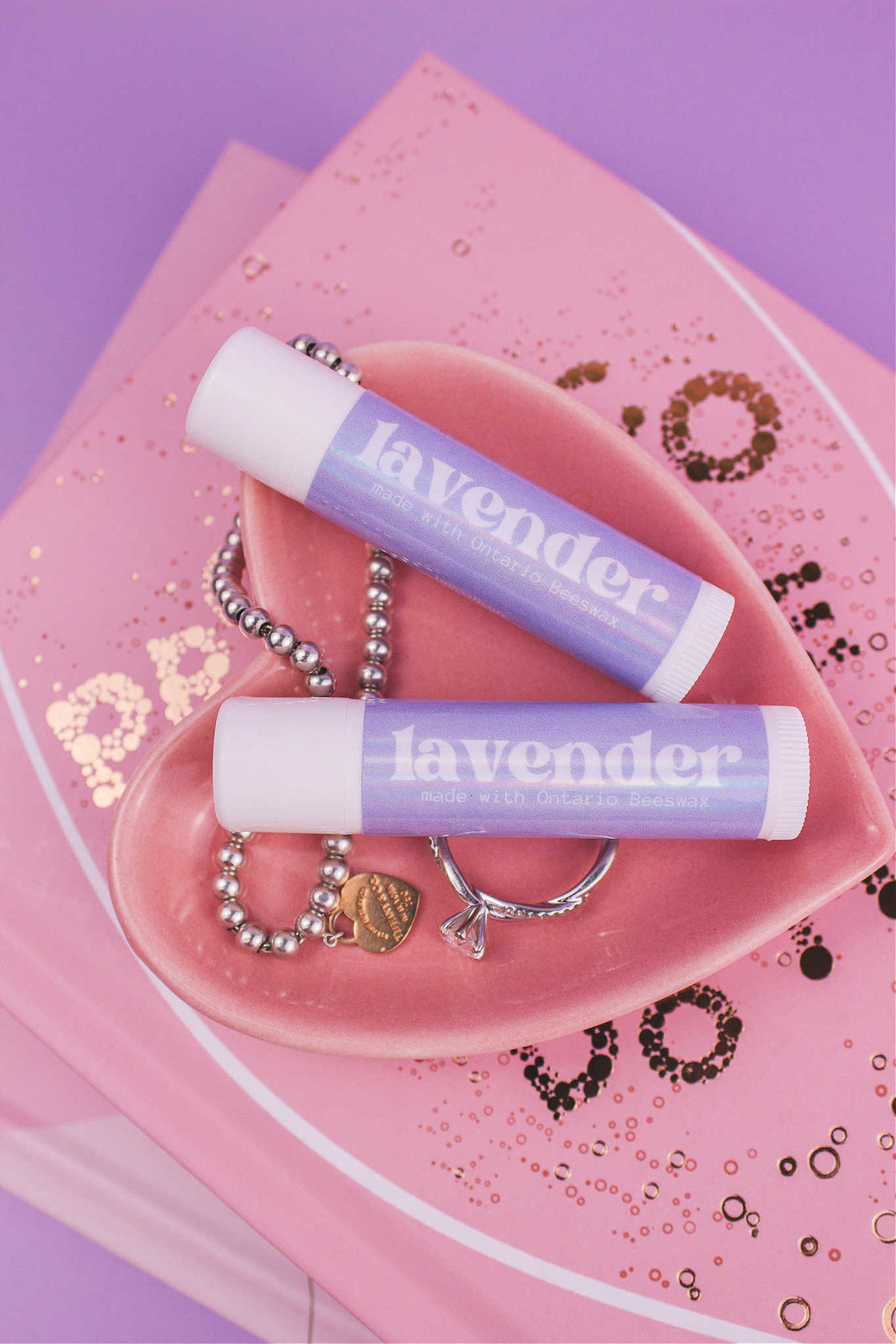 Two Lavender flavoured lip balms displayed in a jewelry dish with a ring and bracelet, sitting on top of books.