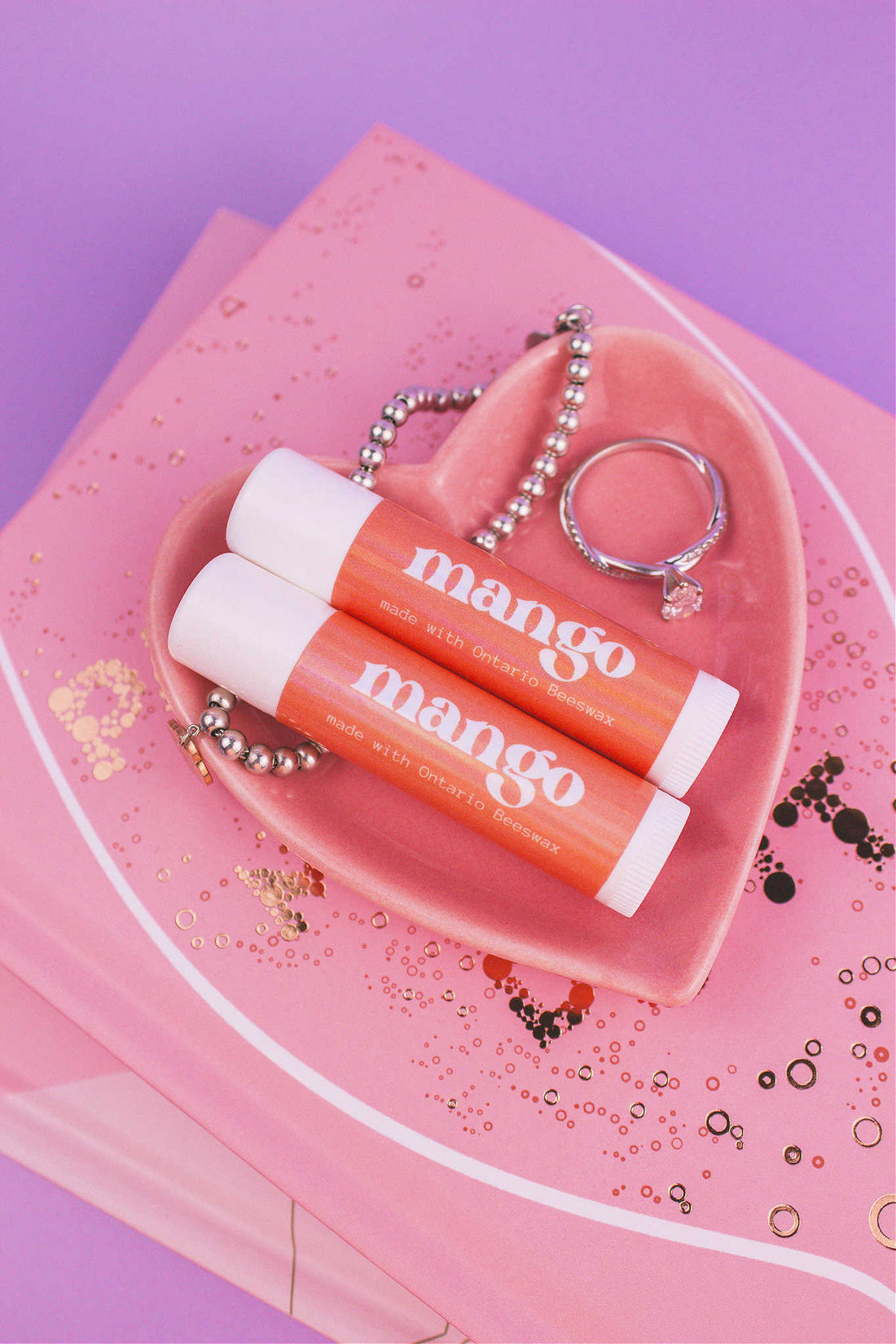 Two Mango flavoured lip balms displayed in a jewelry dish with a ring and bracelet, sitting on top of books.