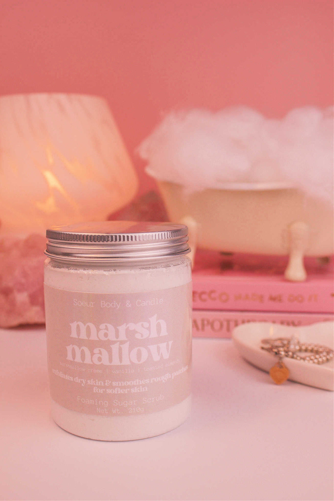 Marshmallow sugar scrub in the foreground, with a small tub filled with bubbles sitting atop a stack of books alongside a jewelry dish, lamp, and crystals. Fragrance notes: marshmallow creme, vanilla, and toasted almond.