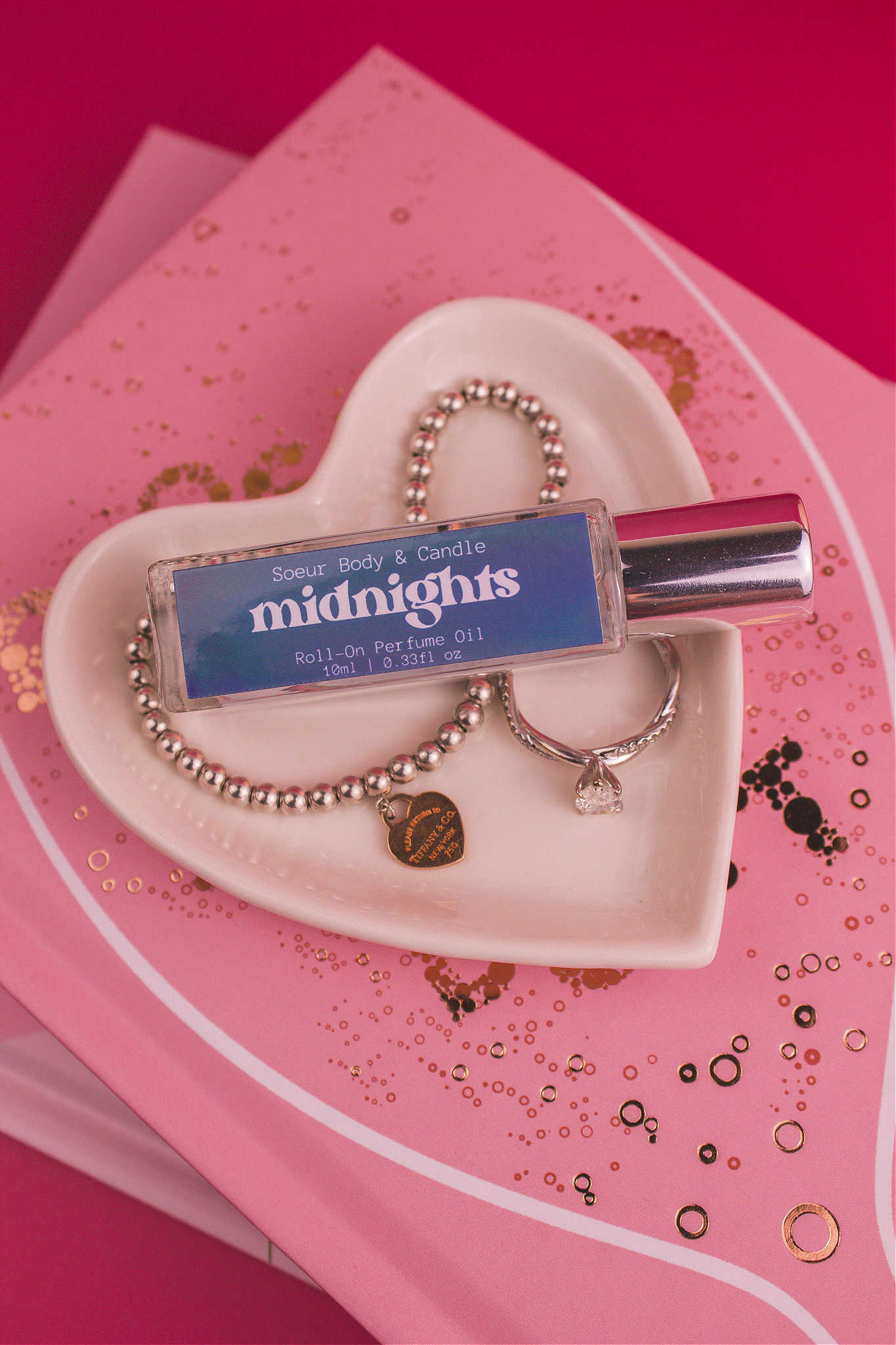 Midnights roll-on perfume bottle in a jewelry dish beside a ring and bracelet, highlighting its daring notes of pear, pepper, jasmine, coffee, and vanilla.