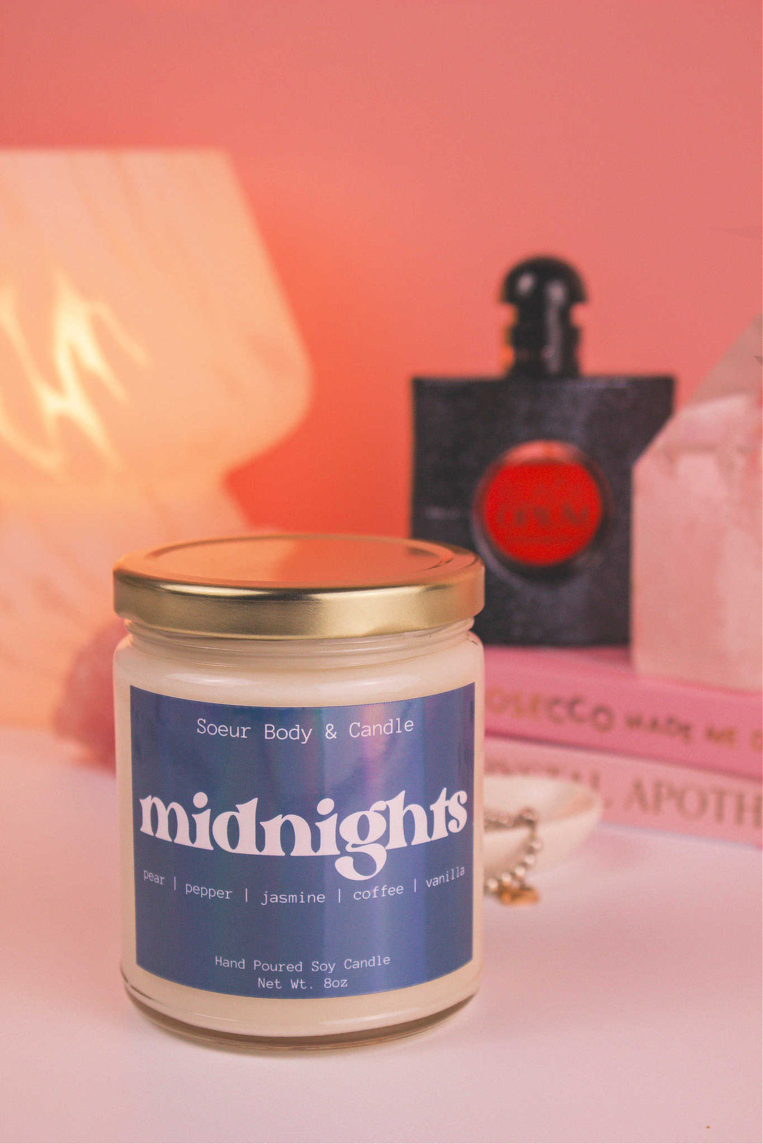 Close-up of Midnights candle with the lid on, with a stack of books, a bottle of perfume, and a jewelry dish in the background, set against a lamp and crystals; fragrance notes: pear, pepper, jasmine, coffee, and vanilla.