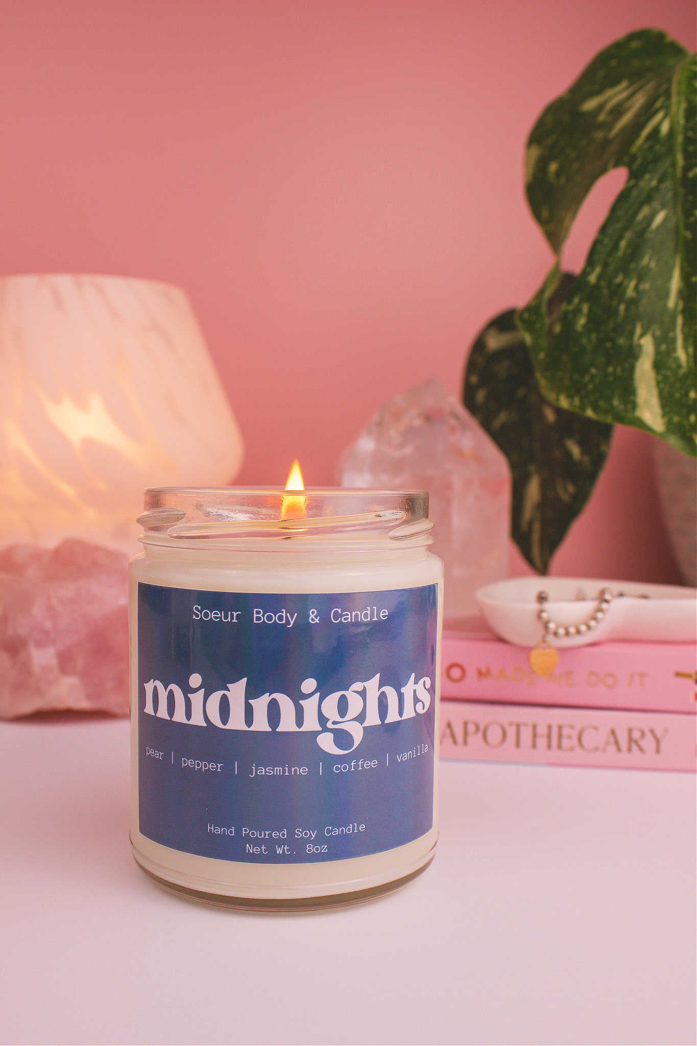 Midnights candle lit, with a stack of books and a jewelry dish in the background, set against a lamp, plant, and crystals; fragrance notes: pear, pepper, jasmine, coffee, and vanilla.